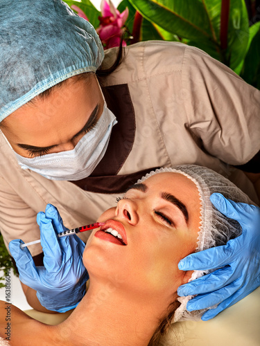 Filler injection for female lips augmentation. Face plastic aesthetic facial surgery in beauty clinic. Doctor in medical gloves with syringe injects lips augmentation. Green plants in background. photo