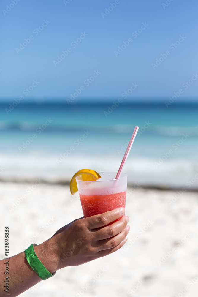 Tanned woman is holding a colorful drink on the beach. Red cocktail with straw and a slice of lemon.