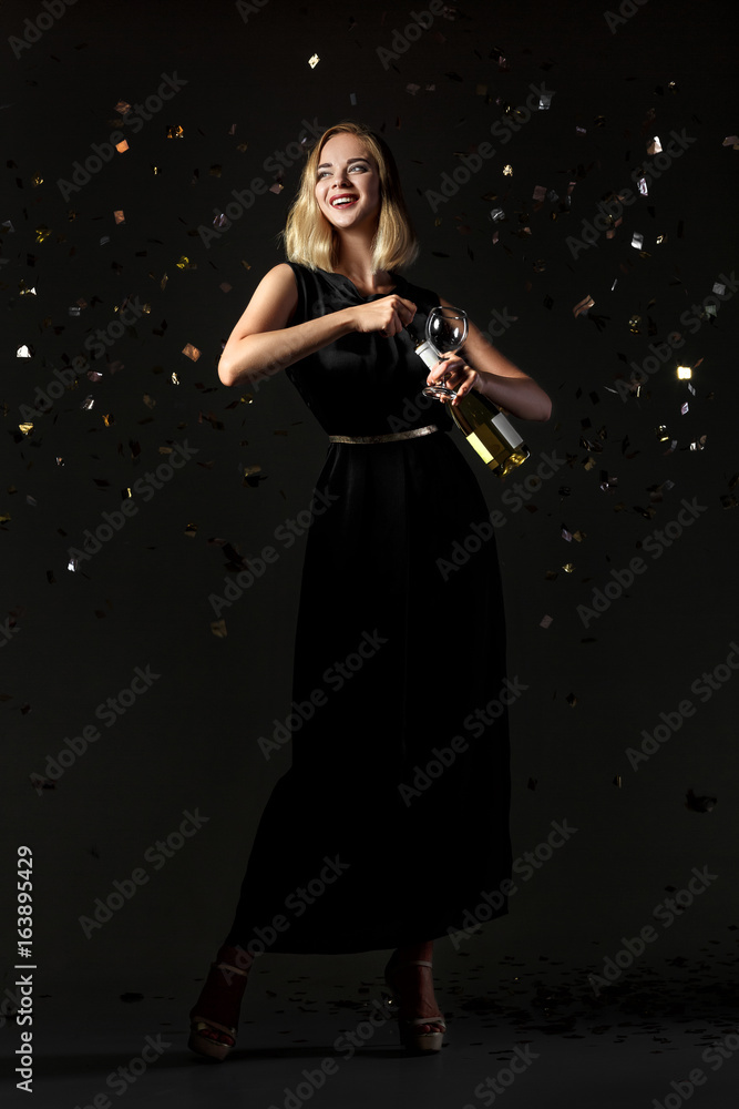 Beautiful happy blonde woman holding glass of white wine on black background with confetti Beautiful happy blonde woman holding glass of white wine on black background with confetti