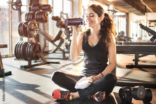 Fitness woman drinking water while sitting and resting on the floor in gym.