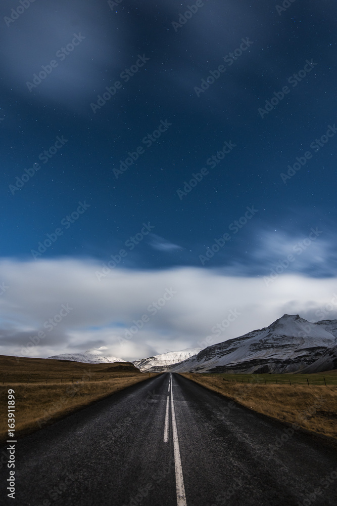 Starry night and empty driving road in Iceland with snowy mountains on the background
