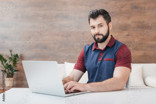 handsome bearded man typing on laptop and looking at camera at home
