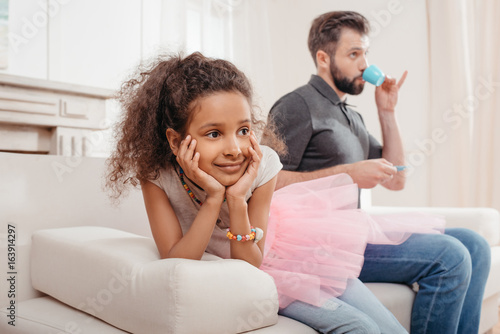 dreamy african american girl sitting on sofa and father drinking from toy cup