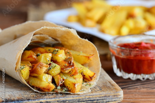 Crunchy fried potatoes with spices. Spicy fried potatoes in paper, ketchup sauce. A wooden background. Quick potato recipe. Closeup