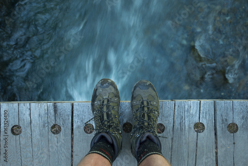 Feet with hiking boots on a wood bridge with flowing water down below