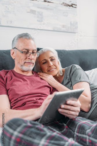 smiling senior couple with digital tablet resting in bed together