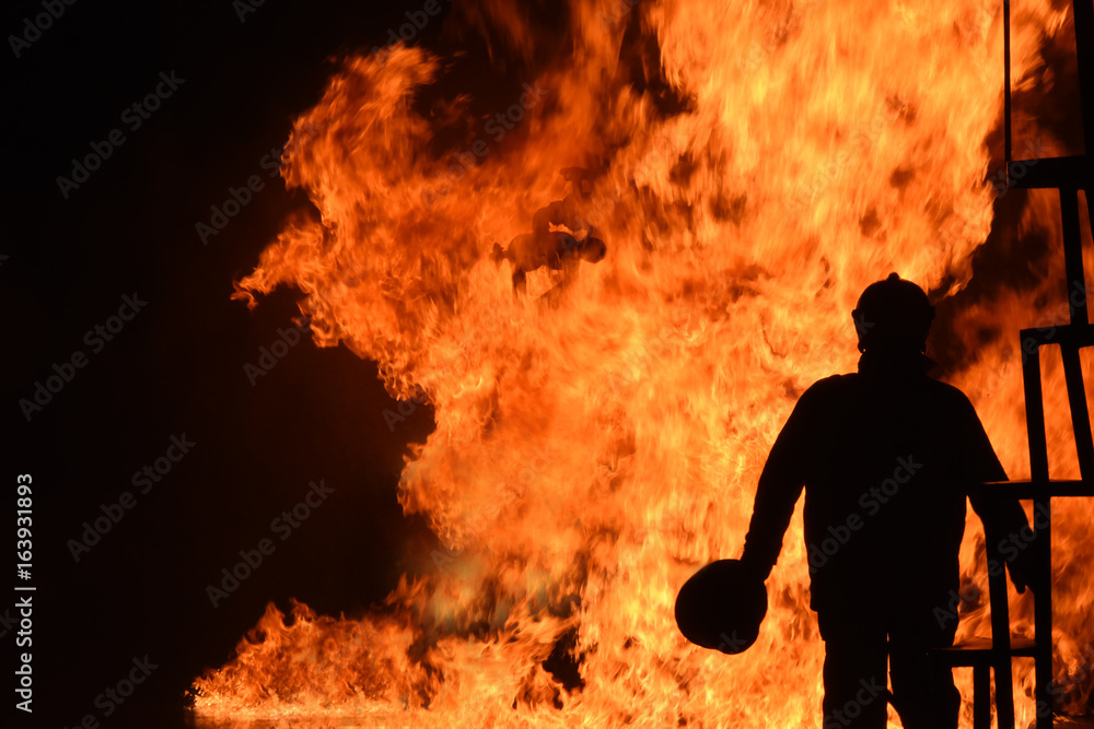 Fototapeta premium Silhouette of Firemen fighting a raging fire with huge flames of burning timber