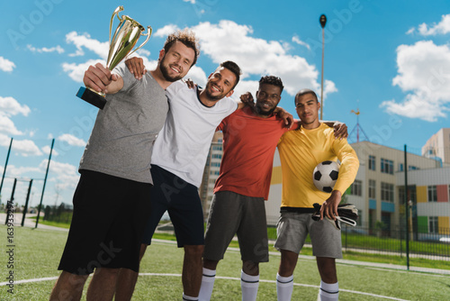 multiethnic soccer team with goblet standing on soccer pitch after game