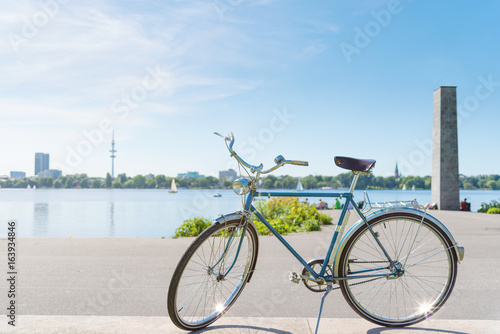 vintage blue bicycle parked at lakeshore of Alster Lake in Hamburg, Germany under beautiful clear blue summer sky with cityscape in blurred background