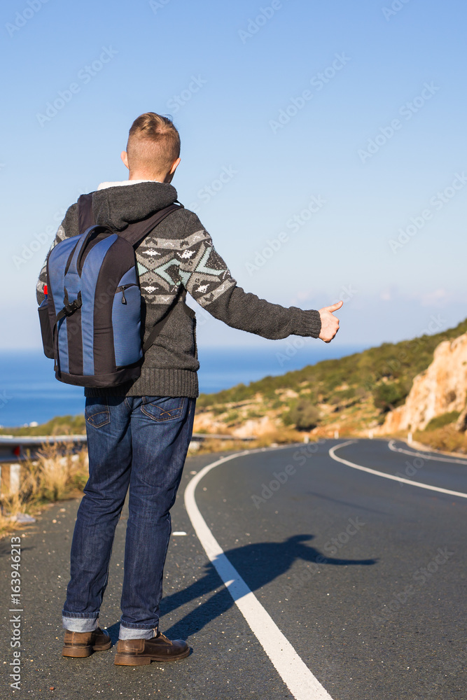 Young caucasian man seen from behind carrying a backpack hitchhiking in a minor road, with his thumb up