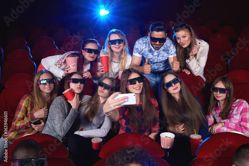 Group of cheerful young friends taking a selfie with a smart phone together while enjoying a 3D movie at the cinema technology gadget social device happiness friendship entertainment leisure concept.