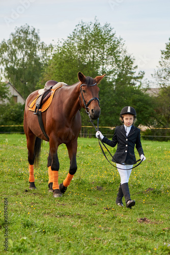 Pretty little girl jockey lead horse by its reins across country in professional outfit © Alex Shadrin