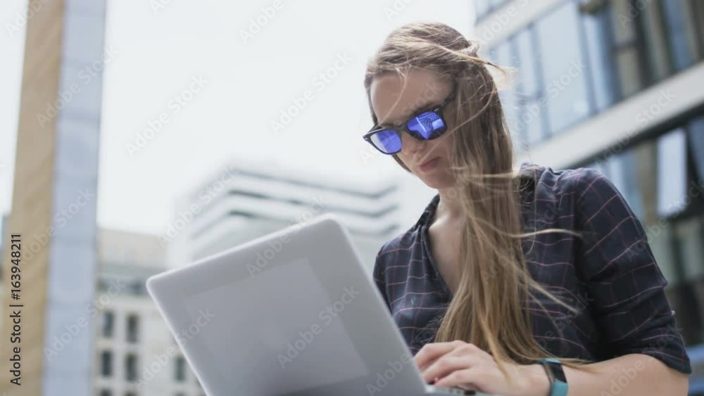 Woman With Dark Glasses Using a Laptop in a Windy City Woman With Dark Glasses Using a Laptop in a Windy City