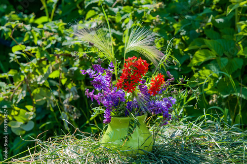 Summer landscape background.A bouquet of purple and red wild flowers in a yellow ceramic vase, standing on dry hay on a background of green leaves.