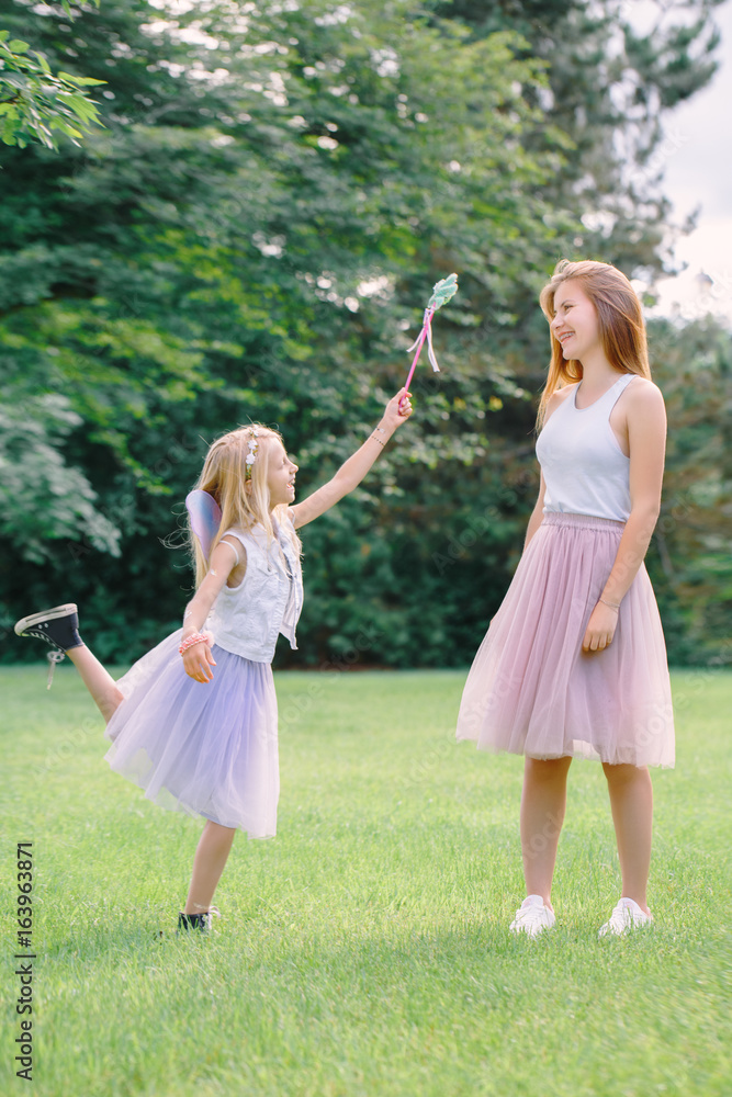 Portrait of two smiling funny Caucasian girls sisters wearing pink tutu tulle skirts in park forest meadow at sunset. Friends having fun together. Girls pretending playing fairies.