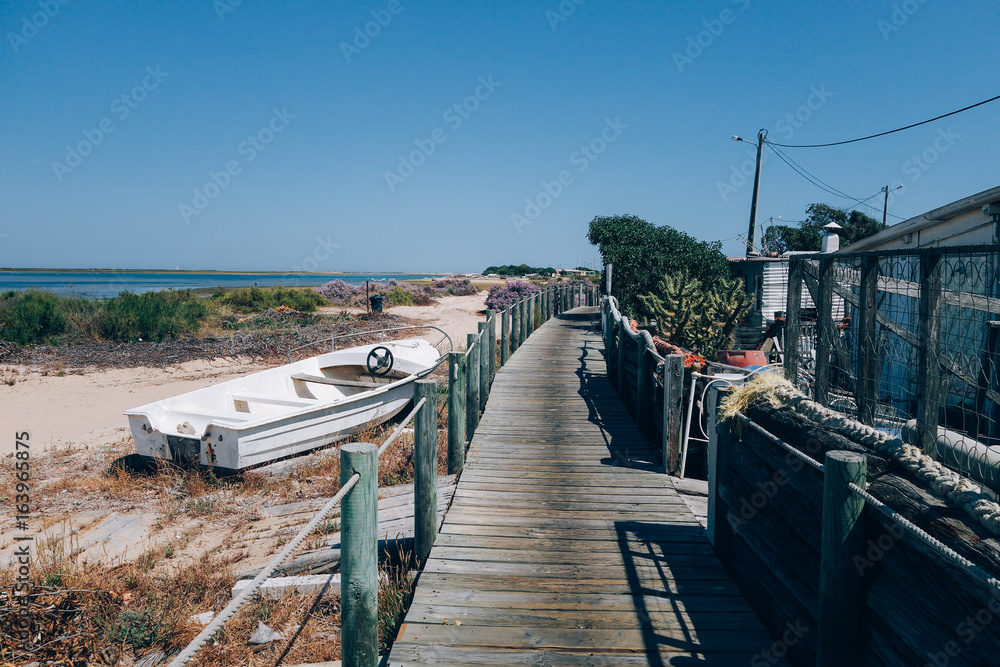 Obraz premium Wooden deck and walk goes into distance on empty beach, with abandoned old fishing motorboats and little huts, hot summer day, unconventional travel destination for nomads