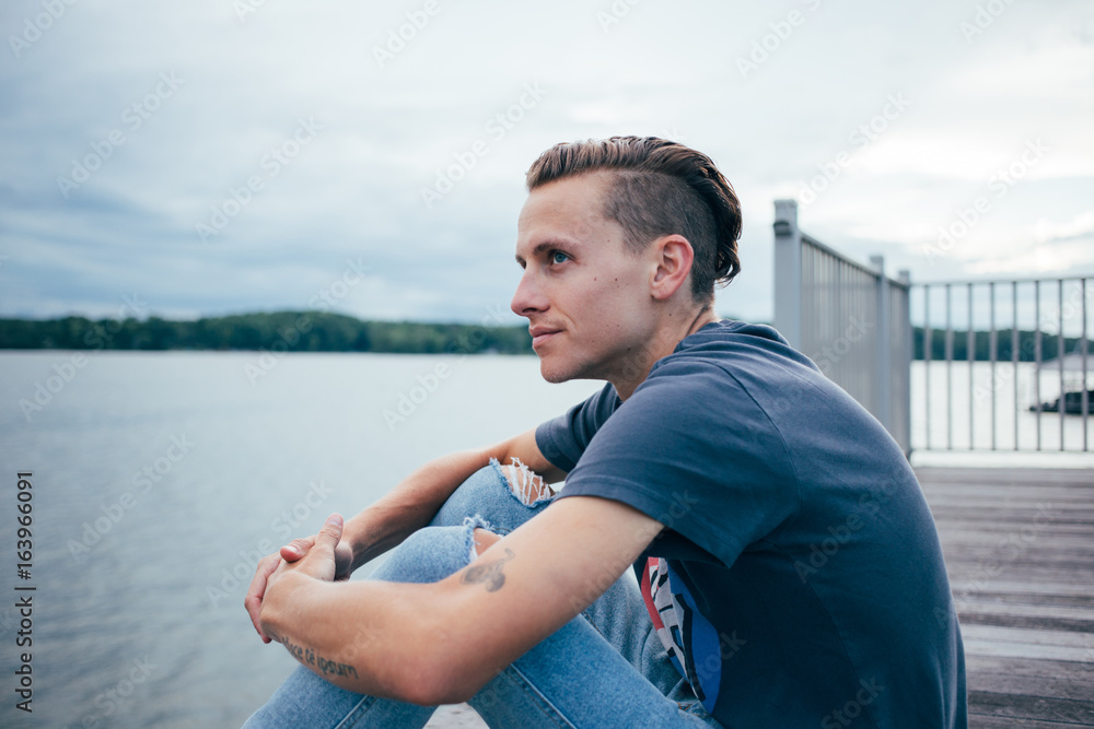 Handsome attractive man sits on deck or dock during sunset waits on boat to come, fashionable and trendy short sides haircut