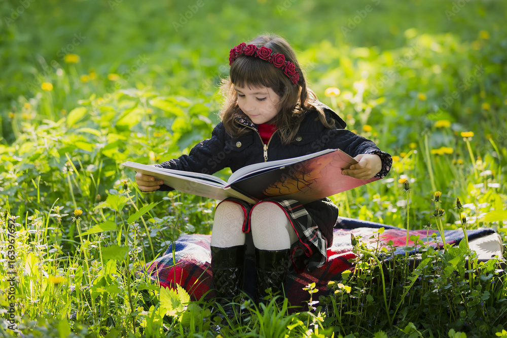 Cute young girl sitting on a suitcase in the grass and reading a book in the park at sunset
