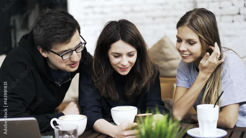 Front view of three young people sitting at a cafe table and watching a video on a smartphone screen. Locked down real time medium shot