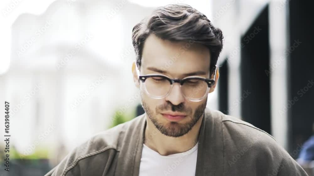 Front view of a young man with a beard and dark hair. He is wearing glasses and a knitted sweater and eating a tasty chunk of meat while listening to his friend. Handheld real time medium shot Front view of a young man with a beard and dark hair. He is wearing glasses and a knitted sweater and eating a tasty chunk of meat while listening to his friend. Handheld real time medium shot