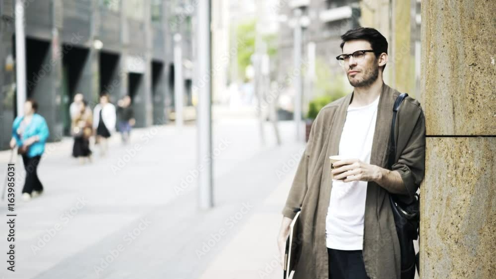 Portrait of a handsome young guy wearing glasses and drinking coffee to go. He is waiting for somebody in the street on a sunny day. Locked down real time medium shot Portrait of a handsome young guy wearing glasses and drinking coffee to go. He is waiting for somebody in the street on a sunny day. Locked down real time medium shot