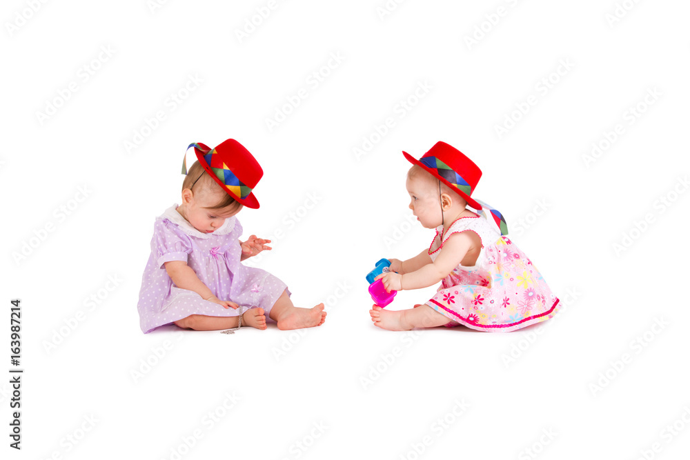 Two lovely babes in colored dresses and red hats are playing on a white background Two lovely babes in colored dresses and red hats are playing on a white background