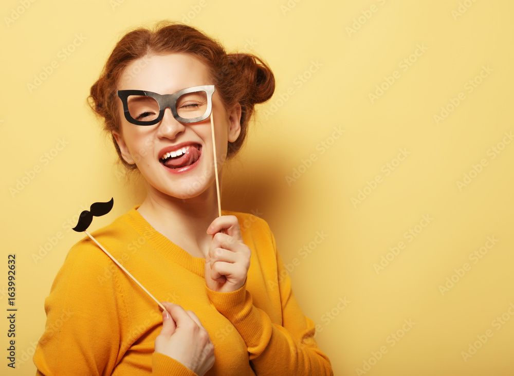 Young woman with fake mustaches and glasses over yellow backgro Young woman with fake mustaches and glasses over yellow backgro