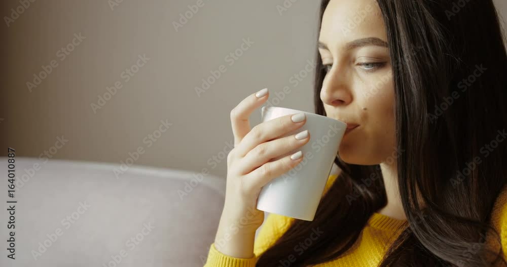 Beautiful young girl sitting on the sofa, drinking coffee, tea and using smart phone. Close up. Slow motion Beautiful young girl sitting on the sofa, drinking coffee, tea and using smart phone. Close up. Slow motion