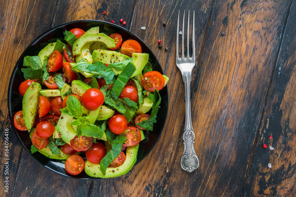 Fotografie Vegetarian salad with avocado, cherry tomatoes and basil leaves on a black plate on the wooden rustic table, top view