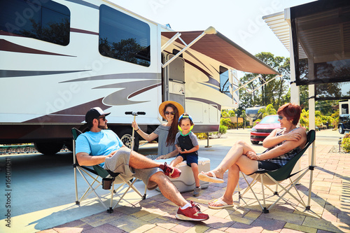 Mother,father,son and grandmother sitting near camping trailer,smiling.Woman,men,kid relaxing on chairs near car.Family spending time together on vacation near sea or ocean in modern rv park photo