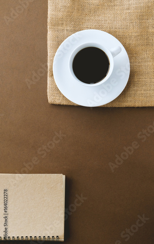top view hot black coffee in a white cup placed on sack and notebook beside on brown background.