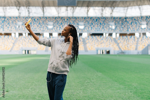 Horizontal photo of the serious afro-american sportgirl taking selfie at the background of the stadium. photo