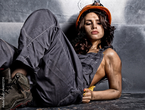Beautiful tired sexy miner worker in orange helmet with flashlight sitting on a floor on steel background photo