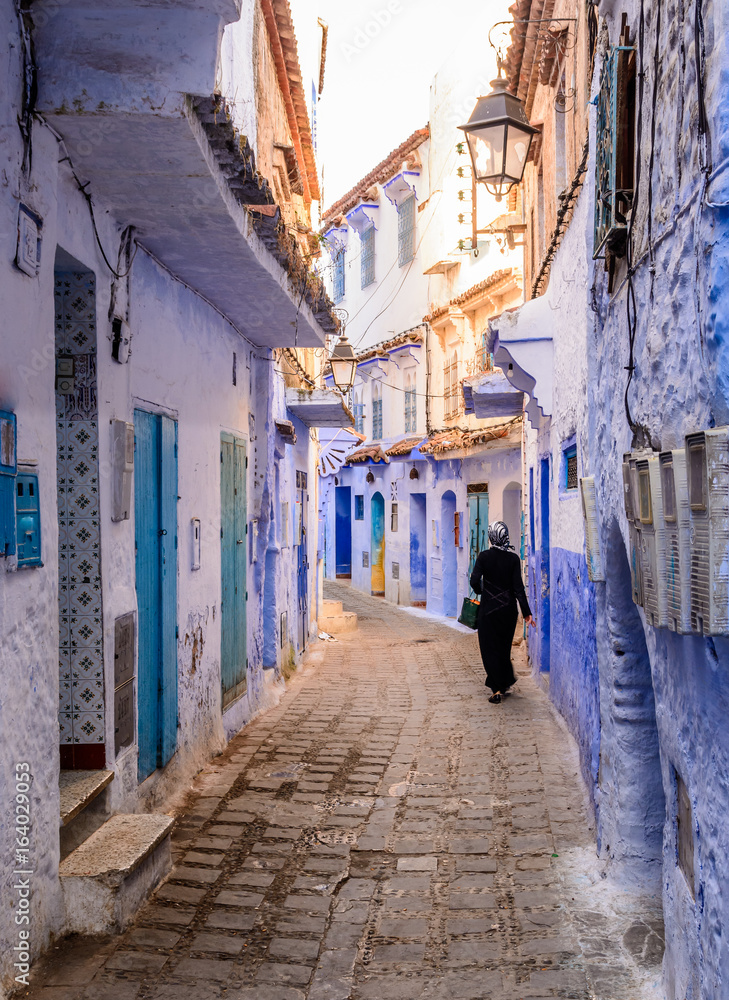 A woman in hijab and black clothing is walking down a street in Chefchaouen, Morocco