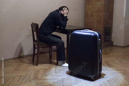 A young man sits bent over the table his head in his hands, .near the table is a large travel bag