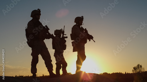 Squad of Three Fully Equipped and Armed Soldiers Standing on Hill in Desert Environment in Sunset Light.