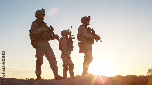 Obraz na plátně Squad of Three Fully Equipped and Armed Soldiers Standing on Hill in Desert Environment in Sunset Light