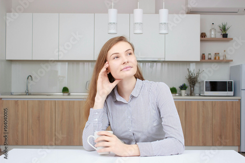 Beautiful cheerful young worker holding coffee cup and looking away with smile talking by phone in morning