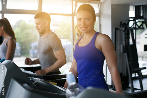 Young man and women workout in gym. Friends together in gym on the treadmill. Woman looking at camera.