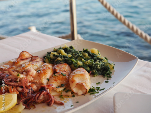 Foto Grilled squid with boiled potatoes and chard (blitva) on white plate with sea on background