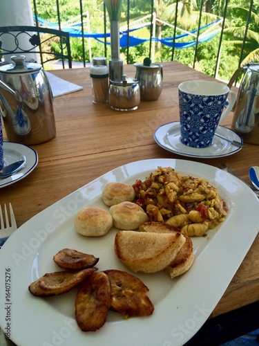 Outside table on the patio with palms: delicious Jamaican breakfast with ackee and saltfish (cod), dumplings, fried banana, plantain, yaam, festival along with fresh hot tea in Port Antonio, Jamaica