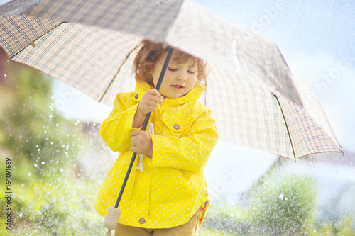 Cute toddler girl wearing yellow waterproof coat and boots with big adult umbrella having fun In the pouring rain. Warm and sunny autumn day