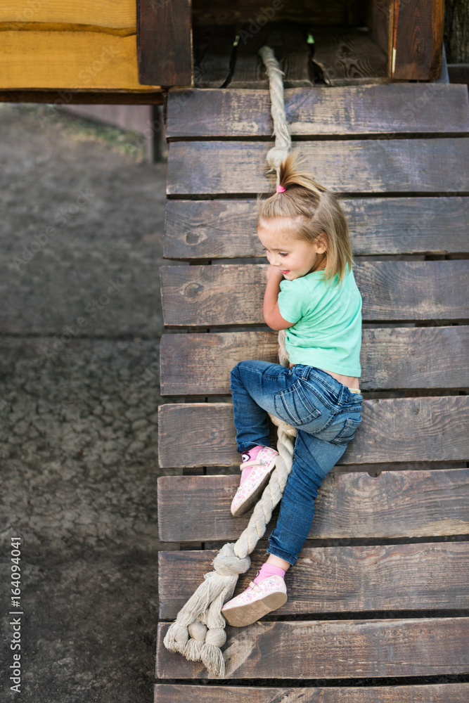Little smiling girl climbs up the rope into the open doorway of a wooden house on the playground Little smiling girl climbs up the rope into the open doorway of a wooden house on the playground
