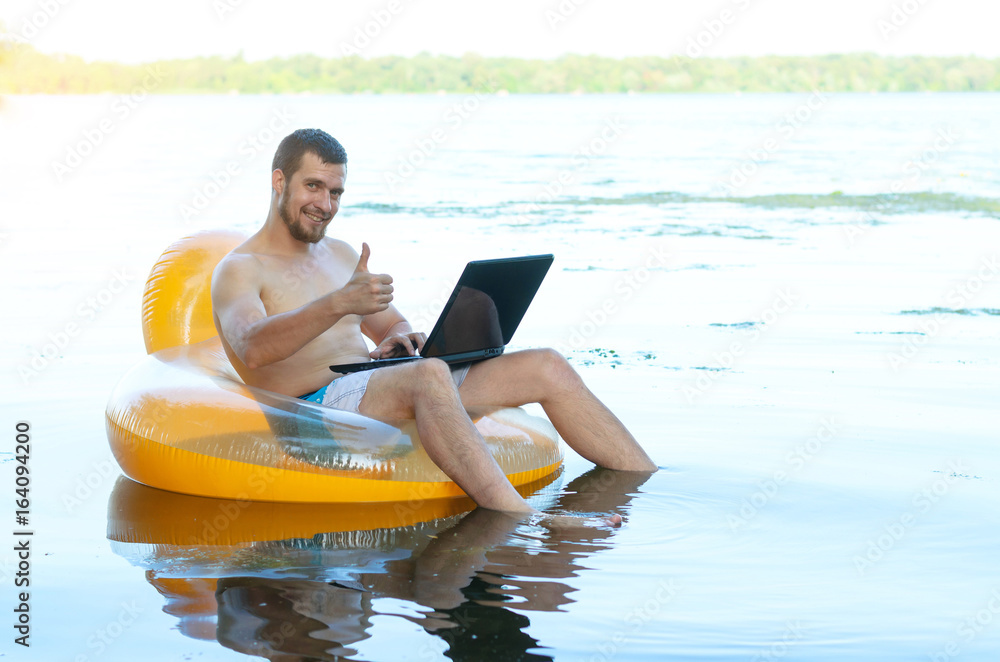 Businessman working on laptop sitting in inflatable ring in water and showing ok sign, free space.