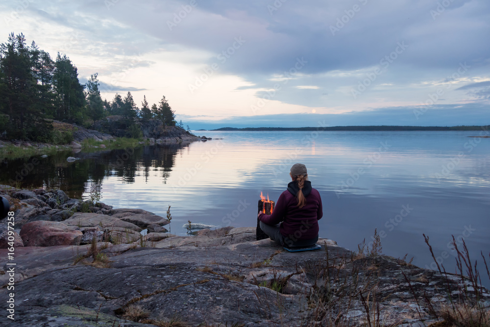 A woman sits by the fire on the stone shore of a large lake. Evening. In the background, you can see a promontory with trees.