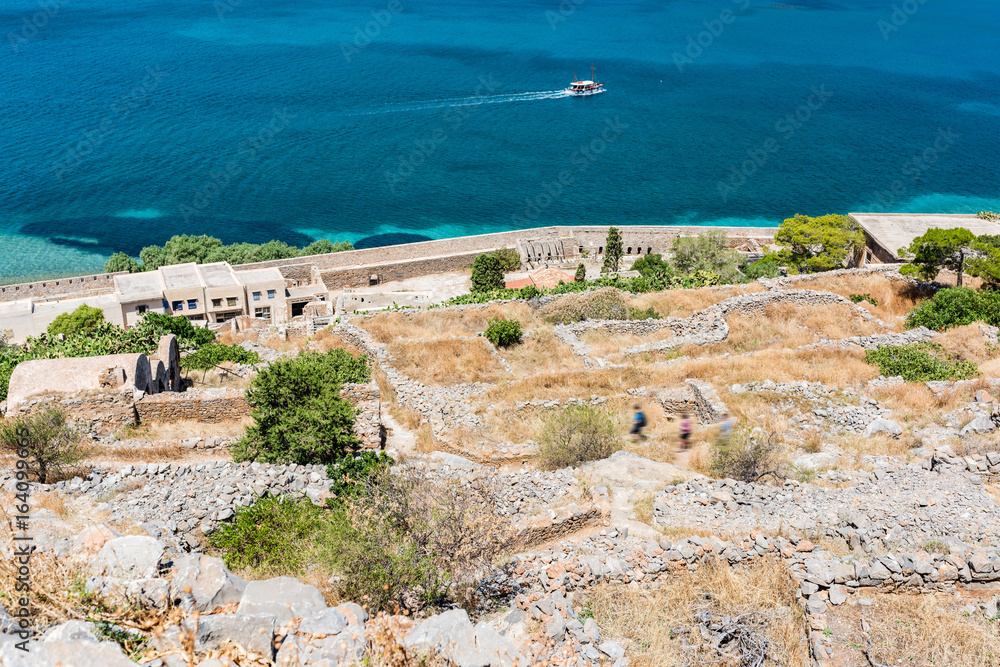 View from top of Spinalonga fortress. Rocky tracks betweenn ruins. Blue sea with boat in front.