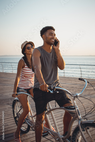 Portrait of a mixed race couple on tandem bicycle outdoors near the sea, young african black man talking on phone © sashafolly