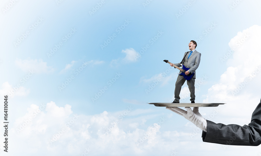 Businessman on metal tray playing electric guitar against blue sky background Businessman on metal tray playing electric guitar against blue sky background