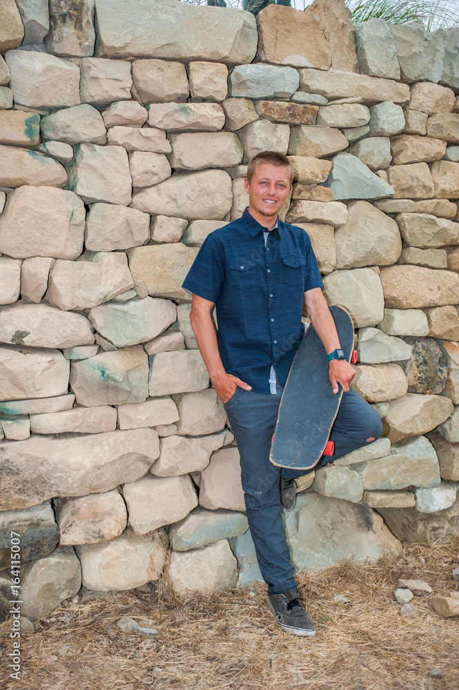 Skater guy leaning against stone wall holding skateboard across thigh in full length view. Skater guy leaning against stone wall holding skateboard across thigh in full length view.