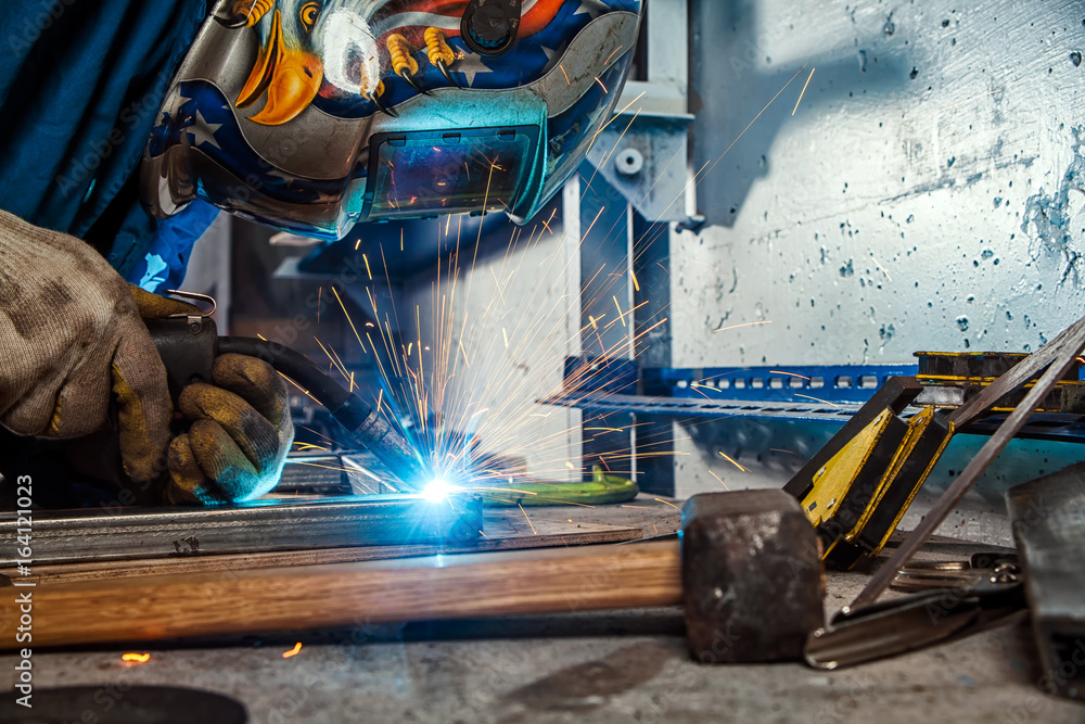 A young man welder in a blue overall, in a welding mask and construction gloves, weld a metal product with a welding machine in the workshop, sparks fly to the sides, close-ups tool on the table